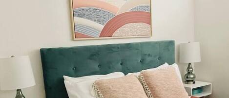 A bed with a green upholstered headboard, two bedside tables with lamps, and a colorful abstract painting above the bed.