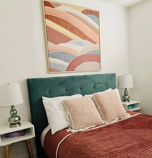 A bed with a green upholstered headboard, two bedside tables with lamps, and a colorful abstract painting above the bed.