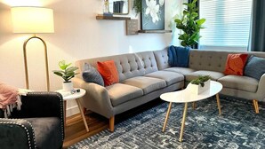 Een moderne woonkamer met een hoekbank, twee bijzettafels, een vloerlamp en een groot raam met zonwering.