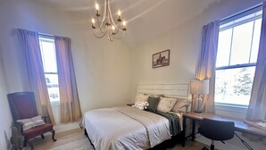 A bedroom with a bed, a chair, a desk, a lamp, and a chandelier. - Opera House Suite 1 (Kemmerer)
