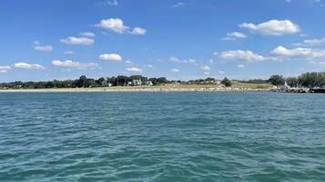 A coastal view with calm waters, a sandy beach, and a line of trees and buildings in the distance.