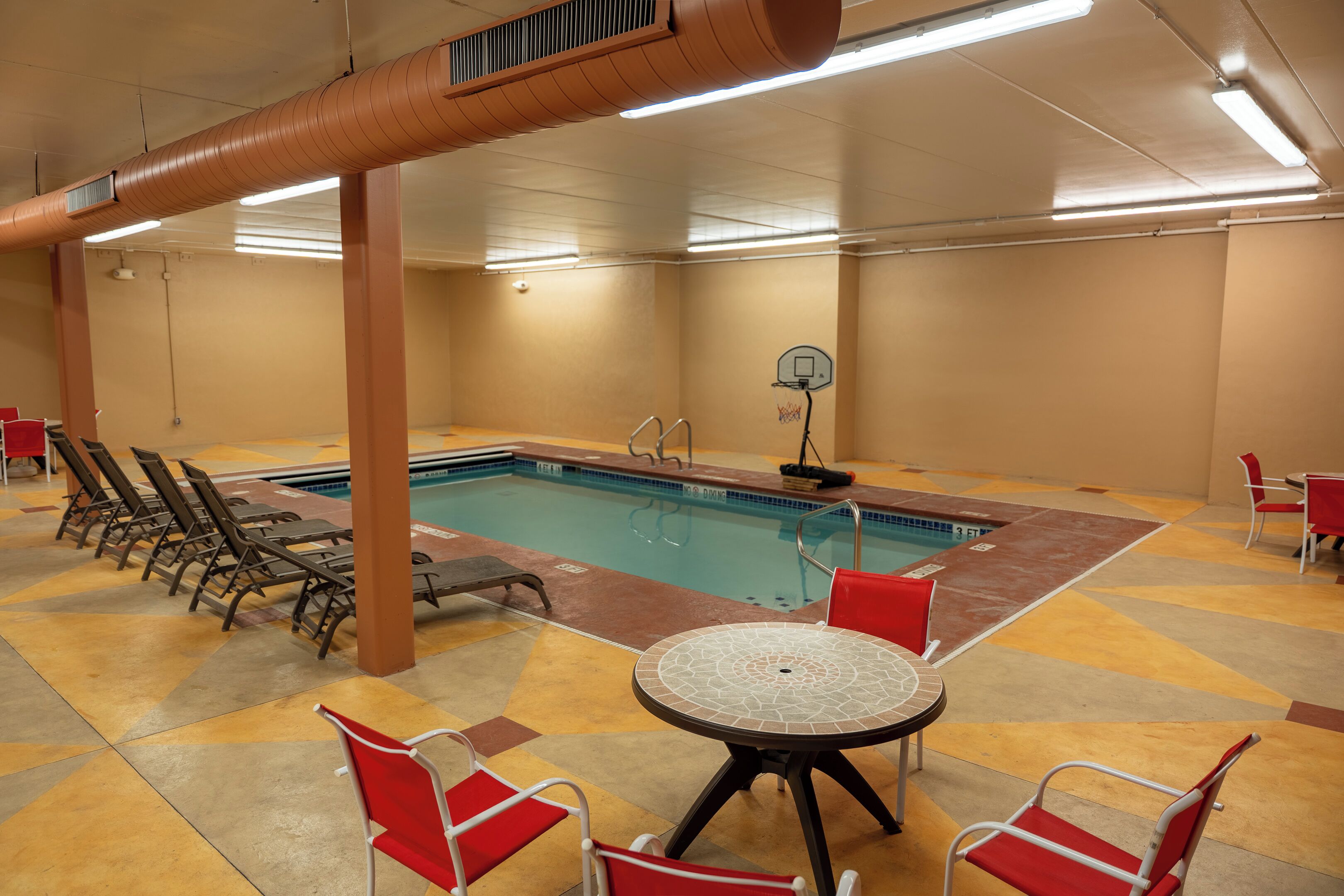 A pool area with lounge chairs and a table, indoor lighting, and a basketball hoop.