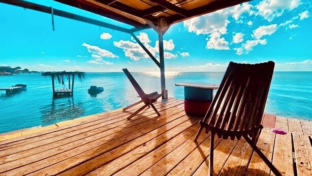 A wooden deck with a chair and a table, overlooking a calm sea with boats and a clear sky. - Enjoy Secret Beach sunsets, fully equipped 3bd house, large deck, AC, WiFi,  (San Pedro)
