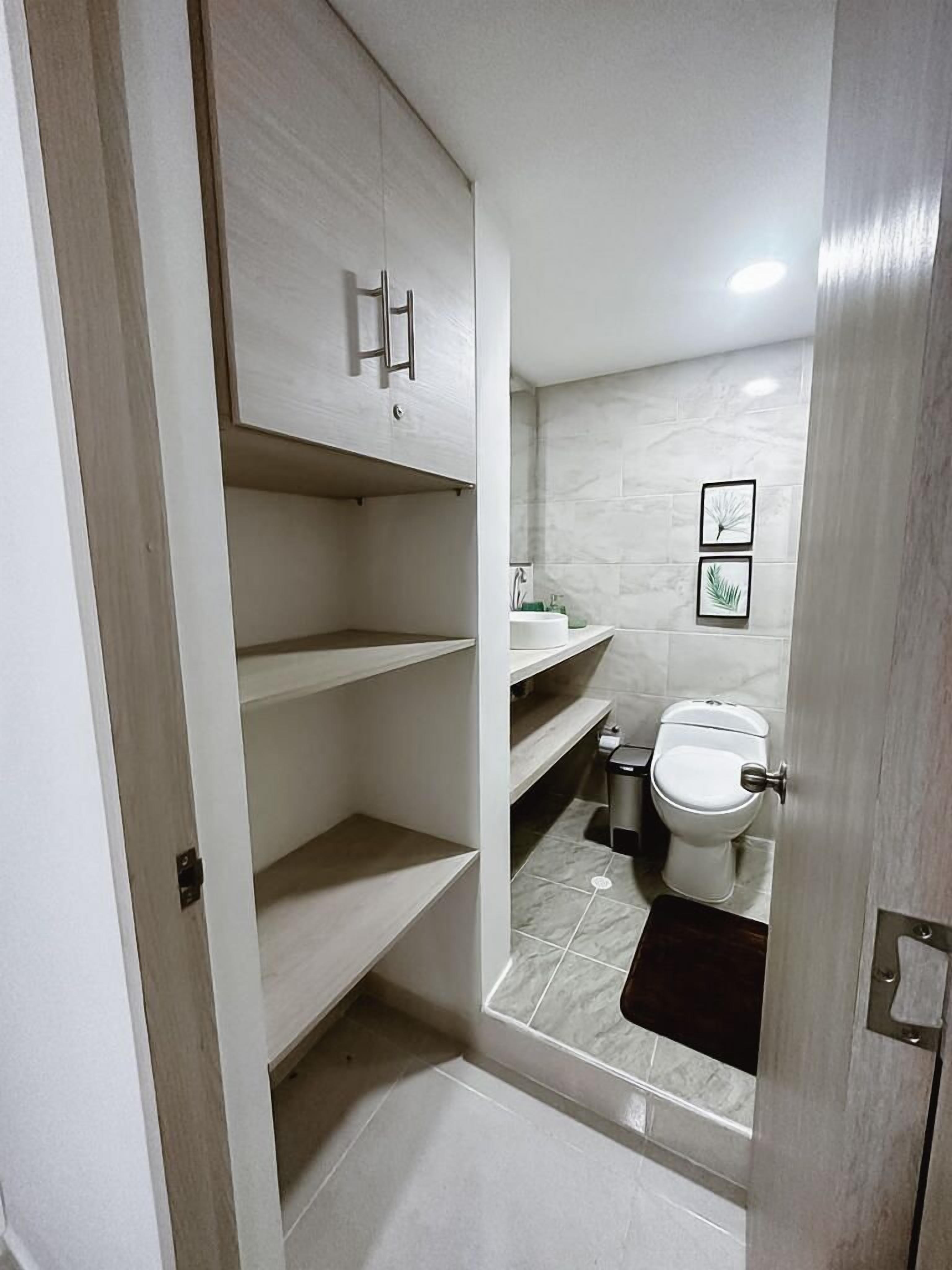 Un bagno con un water, un lavandino e uno specchio. Sopra il lavandino ci sono mensole e una porta che dà su un'altra stanza.