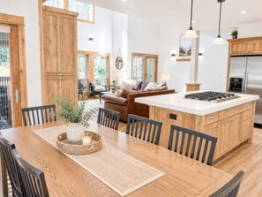 A modern kitchen with a central island, wooden cabinets, and a dining area with a table and chairs. - Serenity Retreat: Near Mt. Bachelor with Pool, Spa, River & Outdoor Trails (Bend)