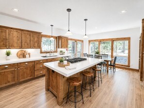 A spacious kitchen with wooden cabinets, a large island with stools, pendant lighting, and large windows offering views of the outdoors.