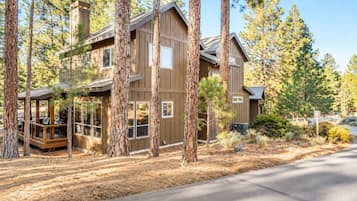 A two-story wooden cabin with a deck and large windows, surrounded by pine trees.