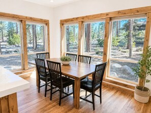 A dining area with a wooden table and chairs, large windows with a forest view, and a potted plant.