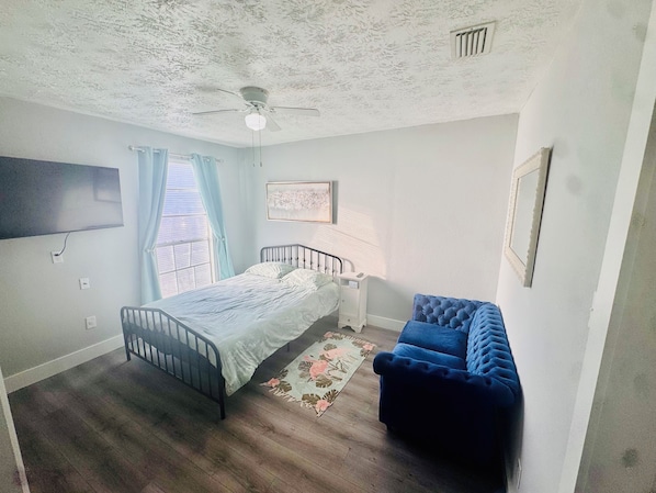 Une chambre à coucher comprenant un lit, une table de chevet, une chaise bleue, un ventilateur de plafond et un téléviseur fixé au mur.