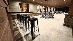 Zona de bar al aire libre con taburetes de madera y pared de piedra, que cuenta con un ventilador de techo e iluminación empotrada.