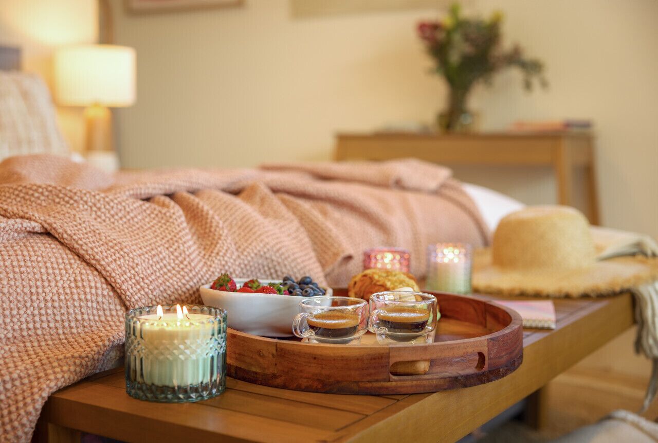 A cozy bedroom setting with a bed covered in a pink blanket, a bedside table with a candle, a bowl of fruit, and two glasses of coffee.