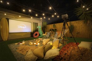 Montaje para una noche de cine al aire libre con pantalla de proyección, un dinosaurio inflable y un área de descanso con cojines y mantas.