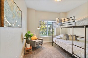 Una habitación con literas, una silla, una mesita pequeña, una planta en maceta y una ventana con persianas.