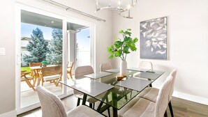 A modern dining area with a glass table, cushioned chairs, a potted plant, and a chandelier. - 2 Adjacent Modern Homes - Sleep 16-20 - Near Shops, Dining, Parks, Entertainment (Meridian)