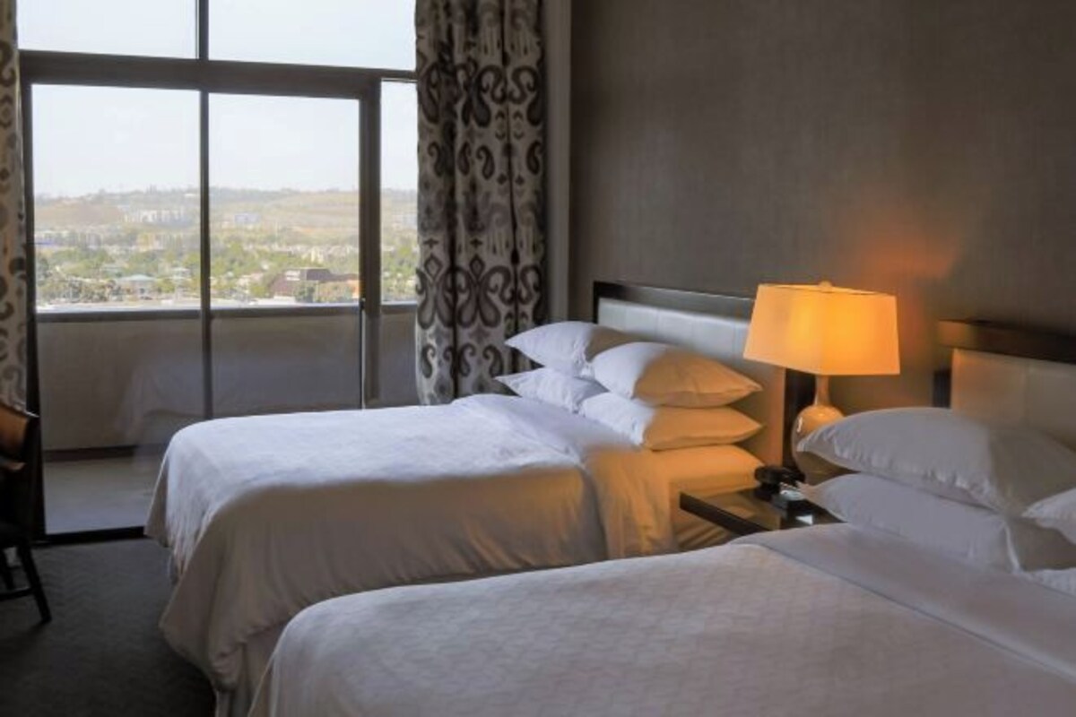 A hotel room with two beds, a nightstand with a lamp, and a view of a cityscape through a large window.