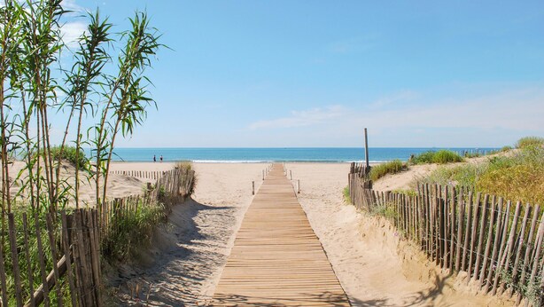 Ein hölzerner Steg führt zu einem Strand bei blauem Himmel.