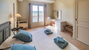 Un dormitorio con cama, mesita de noche con lámpara, ventana, ventilador, espejo y puerta.