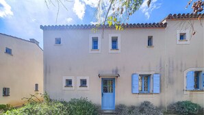 Un edificio de dos pisos con una puerta y contraventanas azules, rodeado de vegetación.