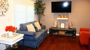 A living room with a blue sofa, a small table with a vase of flowers, a television on the wall, and a red chair. - Cozy, Relaxing 4-Bedroom House in Forest Park (Forest Park)