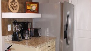 A compact kitchen with a stainless steel refrigerator, a coffee maker, a toaster, and a blender on the counter. There are wooden shelves above the counter holding decorative items and a fire extinguisher mounted on the wall. - Cozy, Relaxing 4-Bedroom House in Forest Park (Forest Park)
