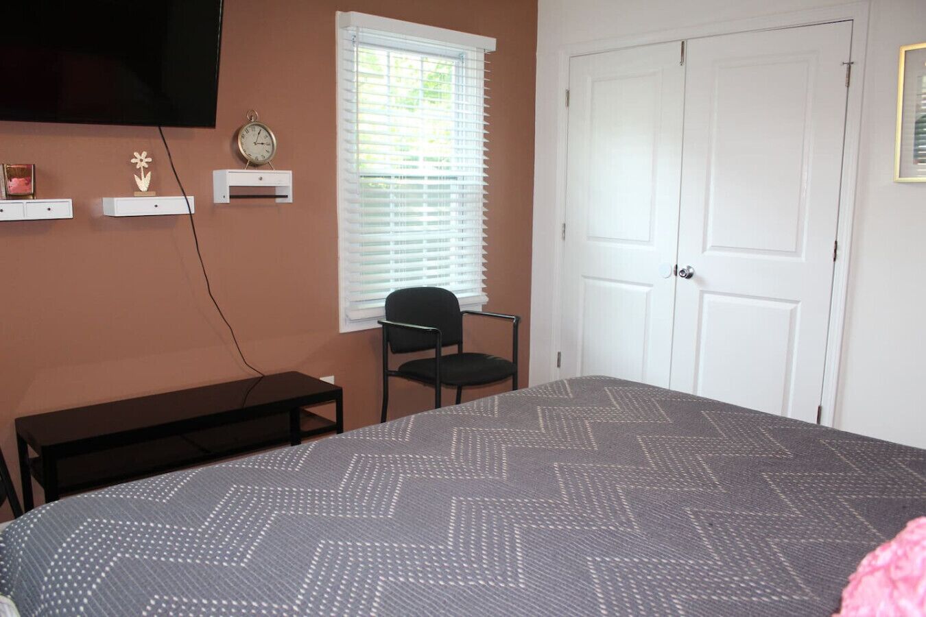 A bedroom with a bed, a chair, a TV, a clock, and a window with blinds.