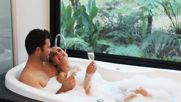 A couple in a bathtub with a glass of champagne, surrounded by lush greenery visible through a window.
