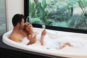A couple in a bathtub with a glass of champagne, surrounded by lush greenery visible through a window. - Ultimate Spa Bower - The Ultimate Spa Bower for a couple (Broulee)