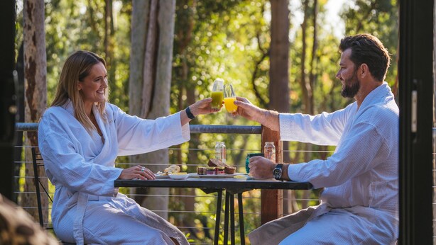 Deux personnes trinquent avec des boissons à une table en plein air, où se trouvent également de la nourriture et des boissons.