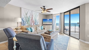 Een woonkamer met een plafondventilator, een bank, een bijzettafeltje en uitzicht op het strand door grote schuifdeuren.