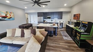 Un salon moderne avec un canapé, une table basse et un baby-foot. On aperçoit une cuisine à l’arrière-plan, avec des meubles sombres et un réfrigérateur.