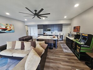 A modern living room with a sofa, a coffee table, and a foosball table. There is a kitchen in the background with dark cabinets and a refrigerator. - Point View 2.0 - Brand New Lakefront Home with Unbeatable Views and Location! (McHenry)