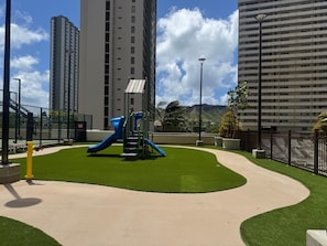 A playground with a slide and climbing structure, surrounded by a well-maintained lawn and tall buildings in the background.