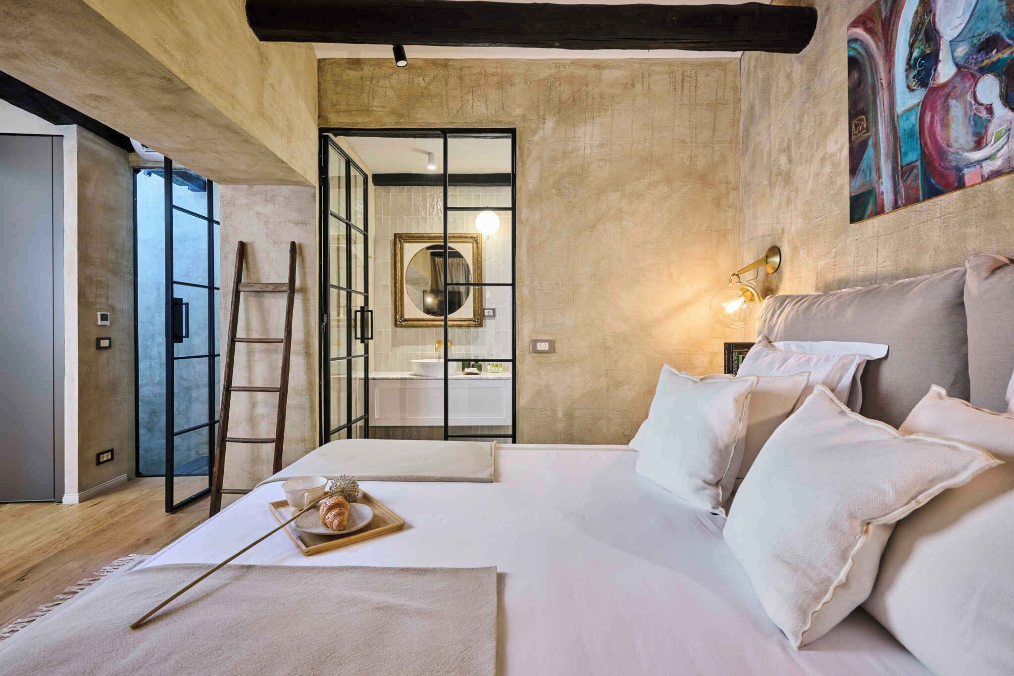 Un dormitorio moderno con una cama grande, una escalera, una bandeja de madera con desayuno, y un baño visible a través de una puerta de cristal.