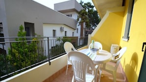 A balcony with white plastic chairs and a small table, a yellow wall, and a view of neighboring buildings. - Luminous Flat With Shared Garden Next to the sea (San Michele al Tagliamento)