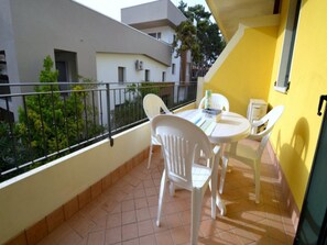 A balcony with white plastic chairs and a small table, a yellow wall, and a view of neighboring buildings and greenery. - Luminous Flat With Shared Garden Next to the sea (San Michele al Tagliamento)
