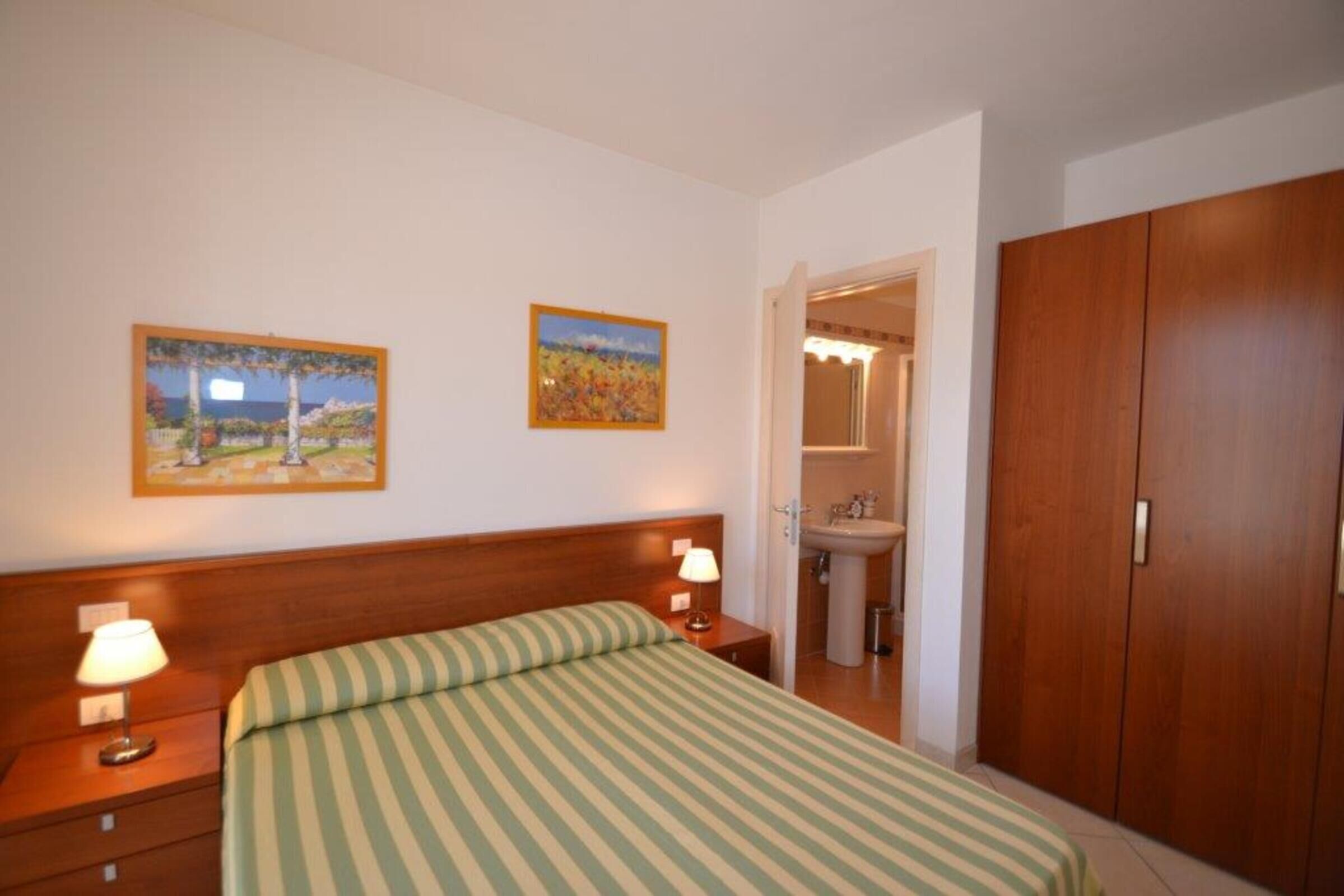 Une chambre à coucher avec un lit, deux lampes de chevet, une armoire en bois et une salle de bain avec un lavabo. Deux tableaux encadrés ornent le mur.