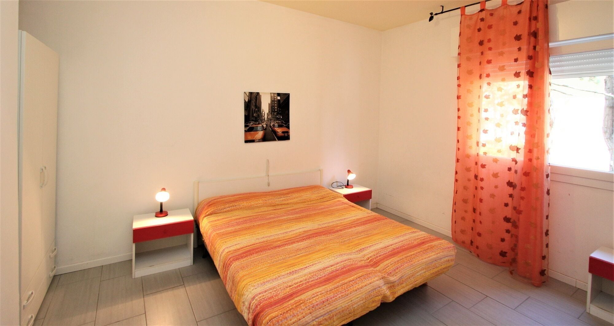 A bedroom with a bed, two bedside tables, a nightstand, a wall-mounted picture, and a window with a curtain.