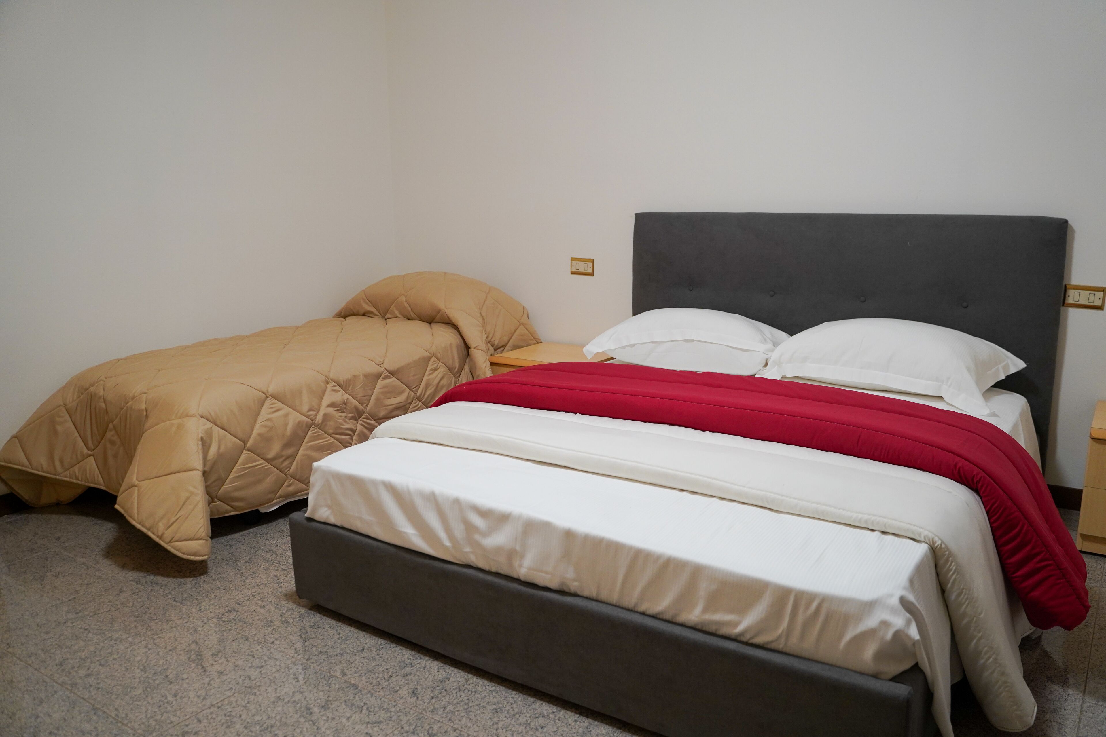 A bedroom with two beds, one with a red blanket and the other with a beige blanket.