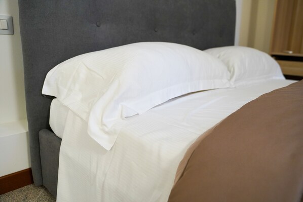 A neatly made bed with white linens and a brown blanket, a grey upholstered headboard, and a wooden nightstand. - Boccadoro Rooms (Mozzecane)