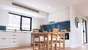 A modern kitchen with white cabinetry, a wooden dining table, and a large window. - Moon Pot at Waters Edge (Mount Martha)