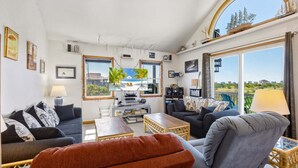 A living room with a sofa, armchair, coffee table, and a TV. There are framed pictures on the wall, a window with a view, and a bookshelf. - 4 br, 2 Min Walk to Beach, hot tub, water view (Avon)