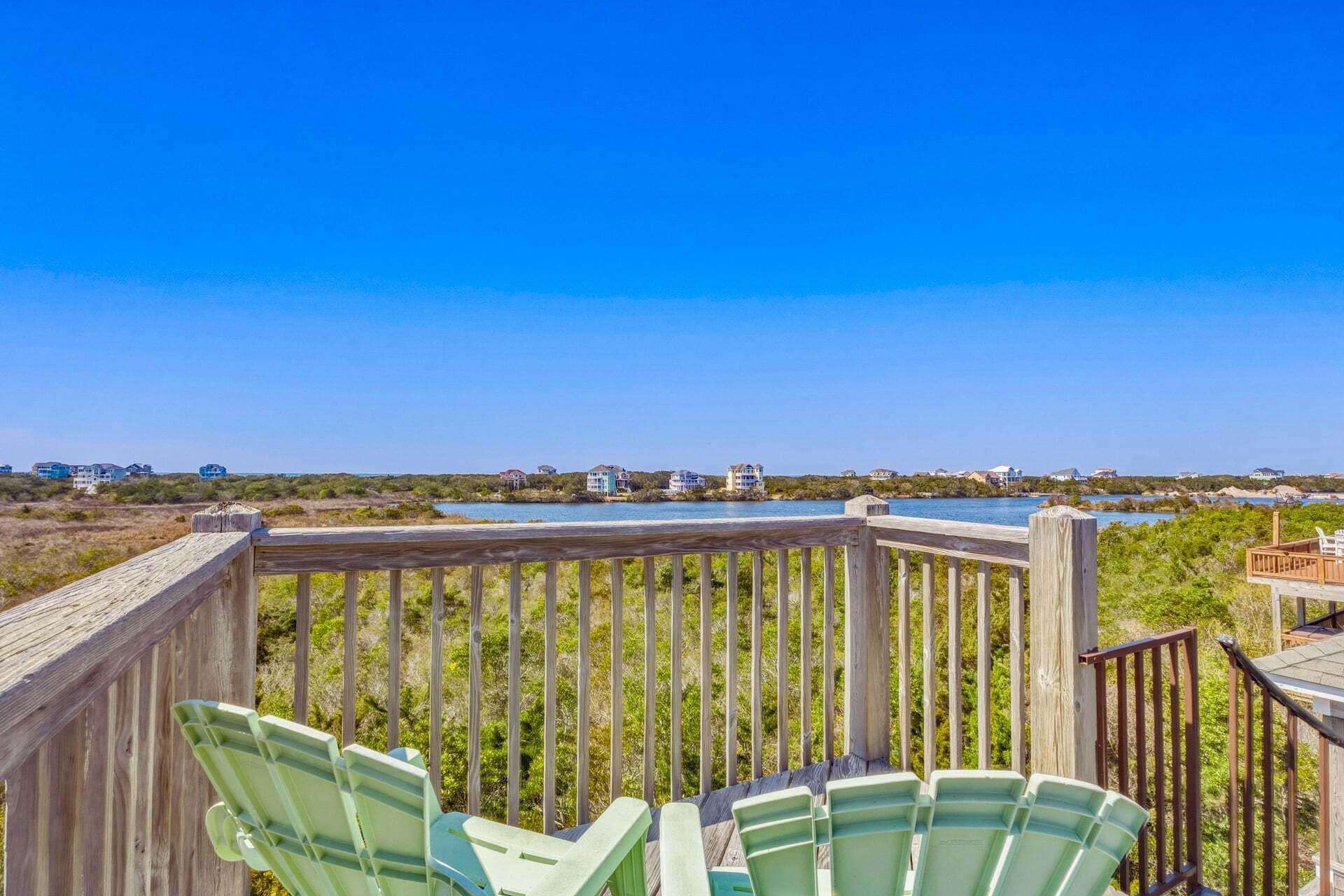 Eine Holzterrasse mit Stühlen, die auf einen Strand und Häuser in der Ferne blickt.