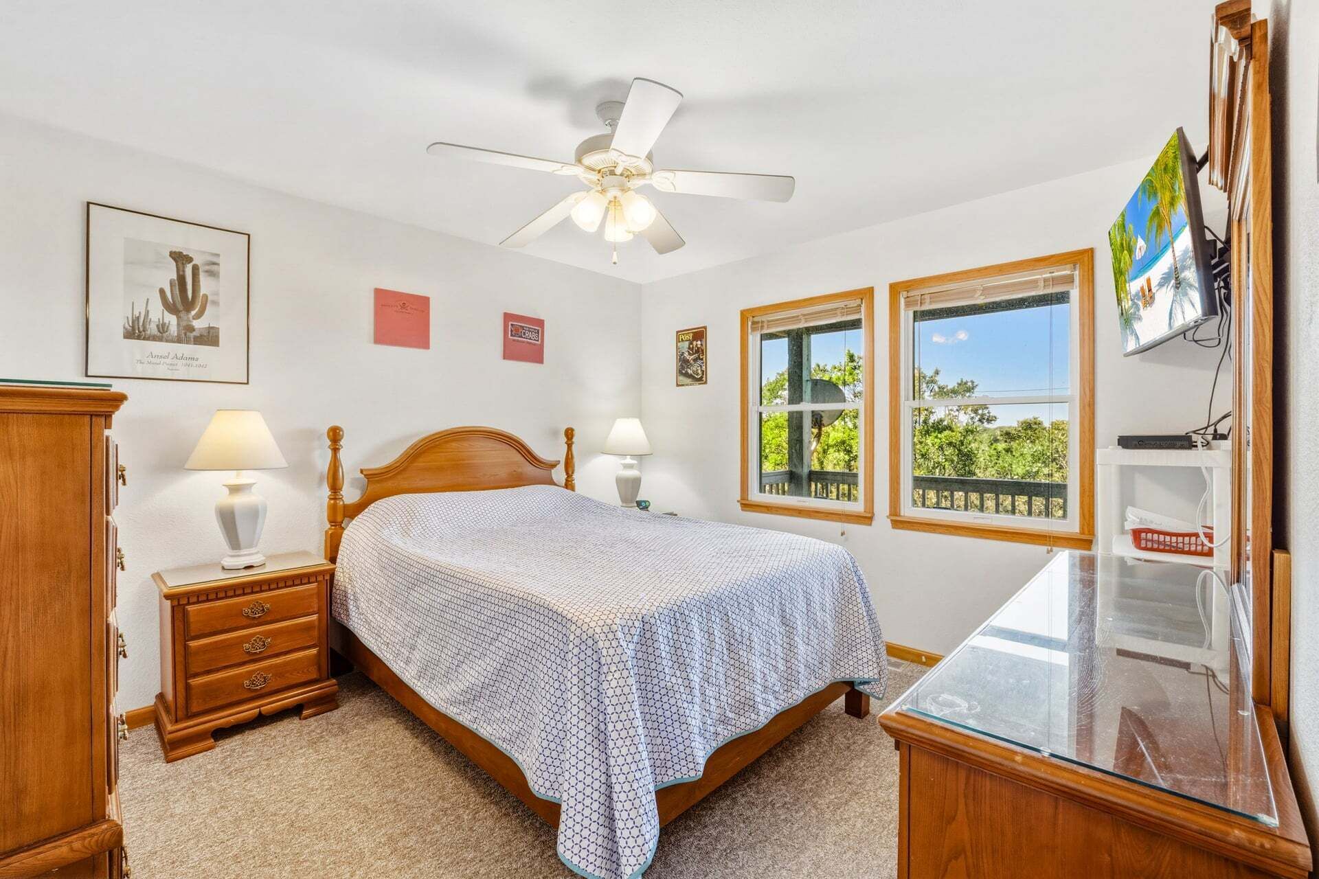 Une chambre à coucher comprenant un lit, des tables de chevet, une commode, un ventilateur de plafond, deux fenêtres donnant sur un beau paysage et des éléments de décoration murale.