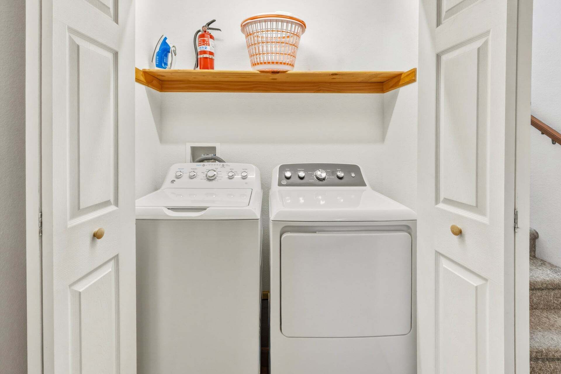 A laundry room with two washing machines, a dryer, and a shelf with a fire extinguisher and a basket.