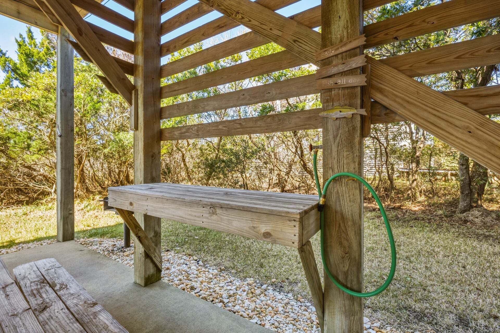 A wooden pergola structure with a bench underneath, surrounded by gravel and trees.
