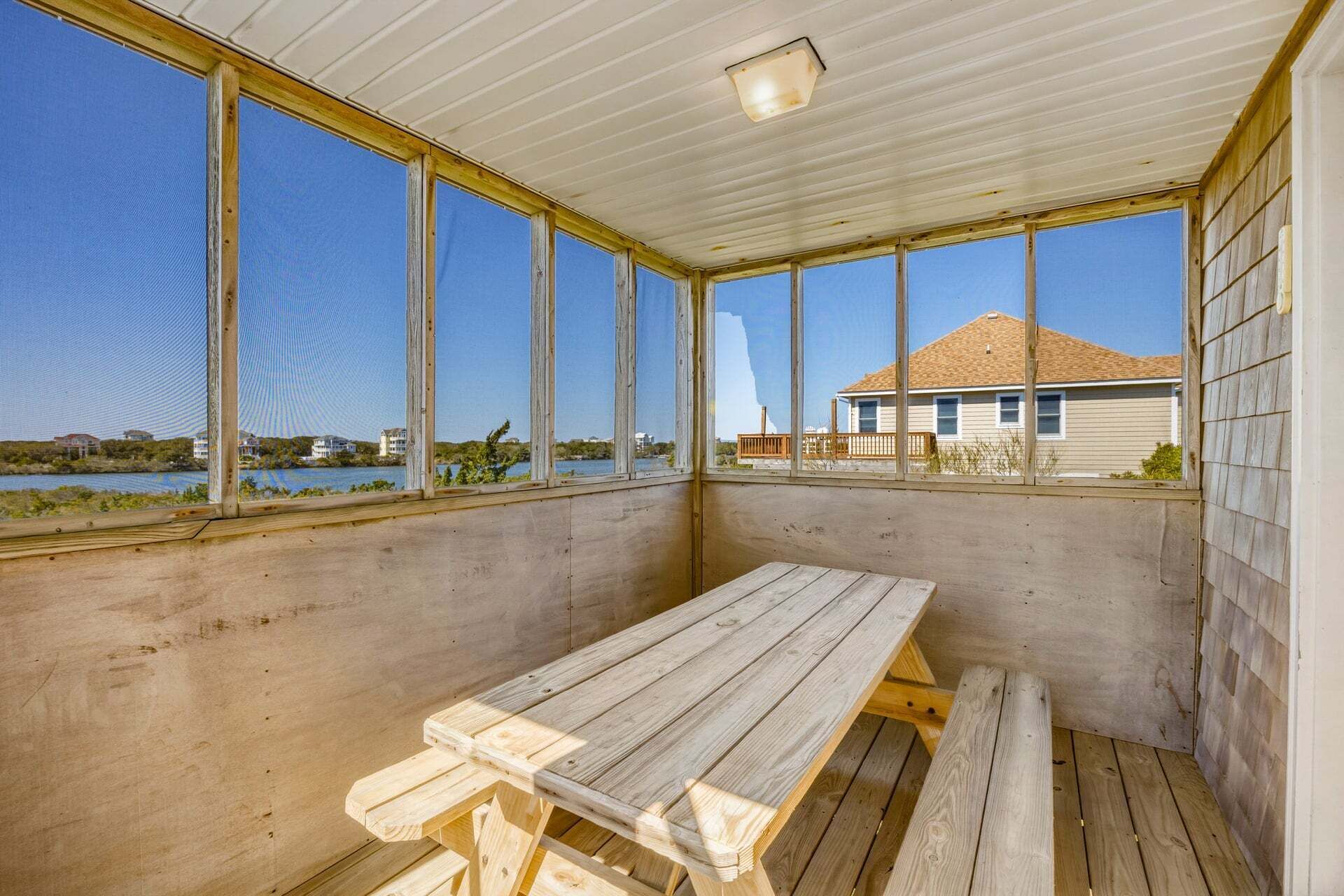A covered outdoor area with a wooden bench, large windows, and a view of a house across a body of water.
