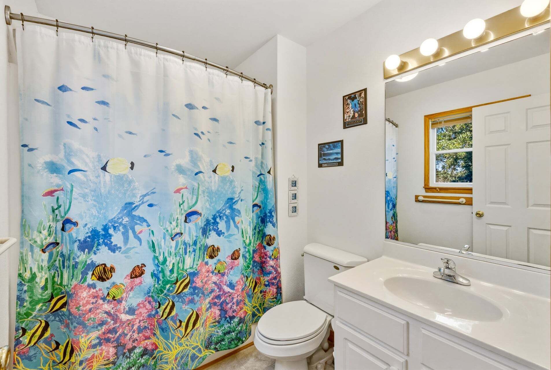 A bathroom with a shower curtain depicting underwater fish and coral, a white toilet, a sink with a mirror, and a window with a view of trees.