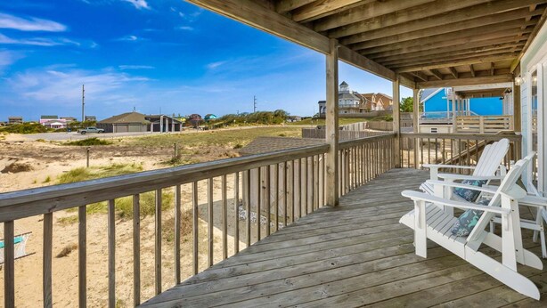 A wooden deck with white chairs, a view of a beach, and houses in the background.