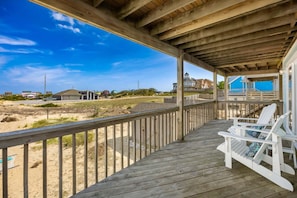 Una terraza de madera con sillas blancas, vista a una playa y casas al fondo.