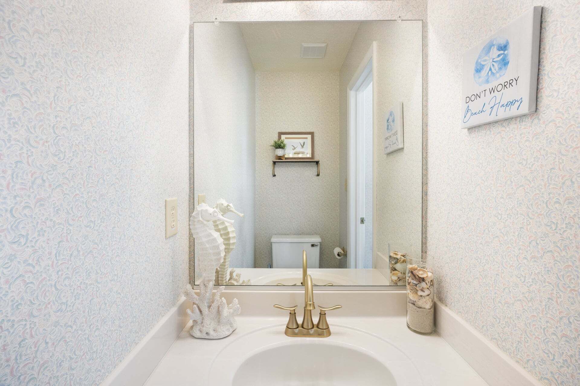 A bathroom with a white sink, gold faucet, and a mirror reflecting a toilet and a framed picture on the wall.
